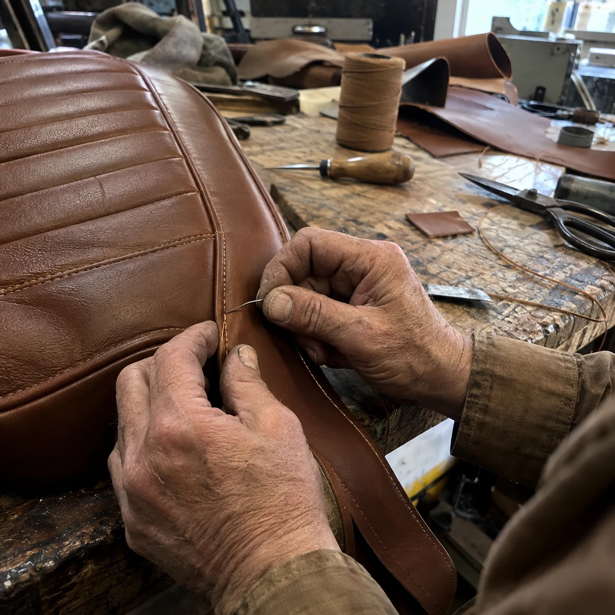 A craftsperson hand-stitching saddle-brown leather upholstery at a worn workbench